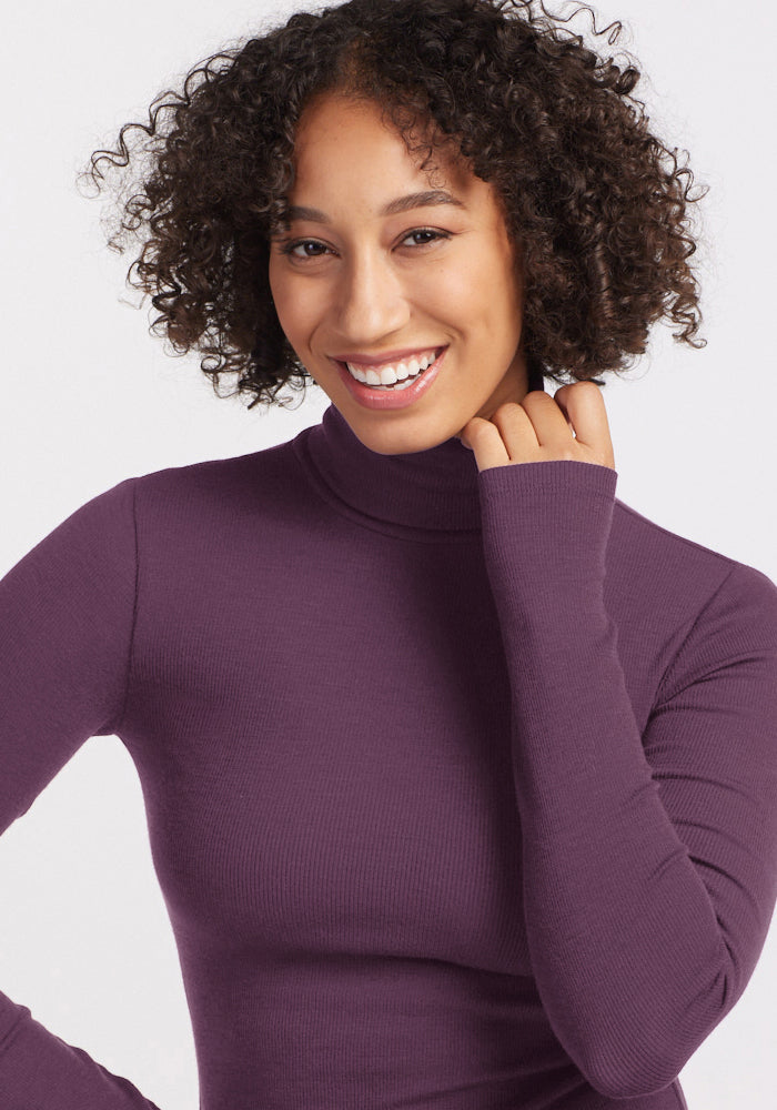 Smiling person with curly hair wears the Woolx Sage Ribbed Turtleneck in Purple Iris against a light background.