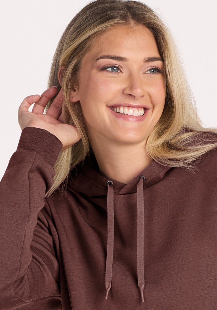 Smiling woman with long blonde hair wearing the Woolx Avery Hoodie in Canyon Clay, hand near her ear.
