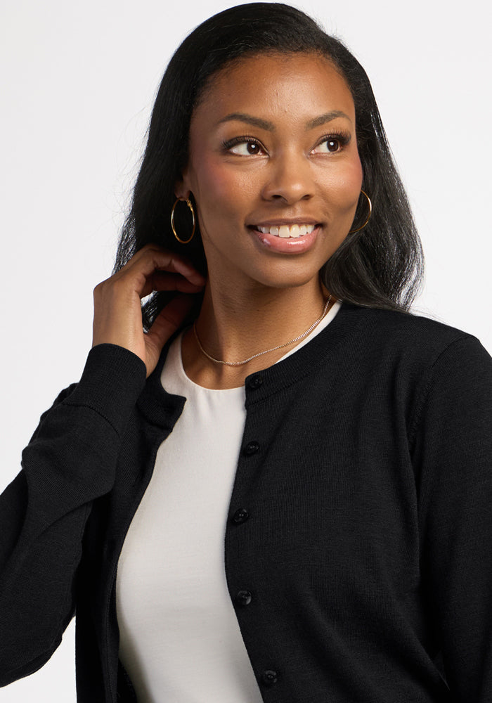 Woman wearing Woolx Caitlin Sweater in black over a white shirt, smiling and looking aside, hand near neck.
