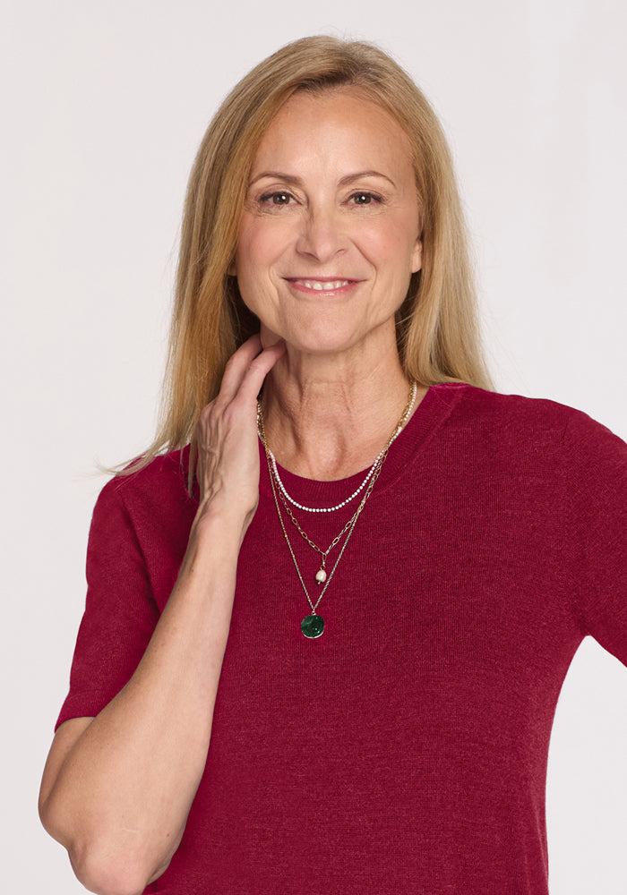 Smiling blonde woman in Woolx Anna Crew Sweater - Beet Red, layered necklaces, poses with hand on her neck.
