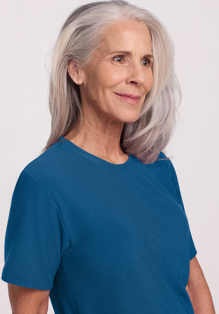 An older woman with long gray hair and a gentle smile wears the Woolx Delia Ribbed Short Sleeve in Moroccan Blue, standing against a light background and gazing slightly to the side.