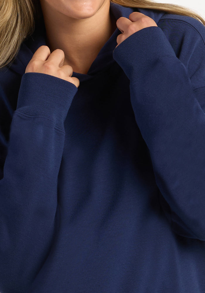 A person pulls the hood of their dark blue Woolx Genevieve Hoodie - Ocean Cavern up with both hands.