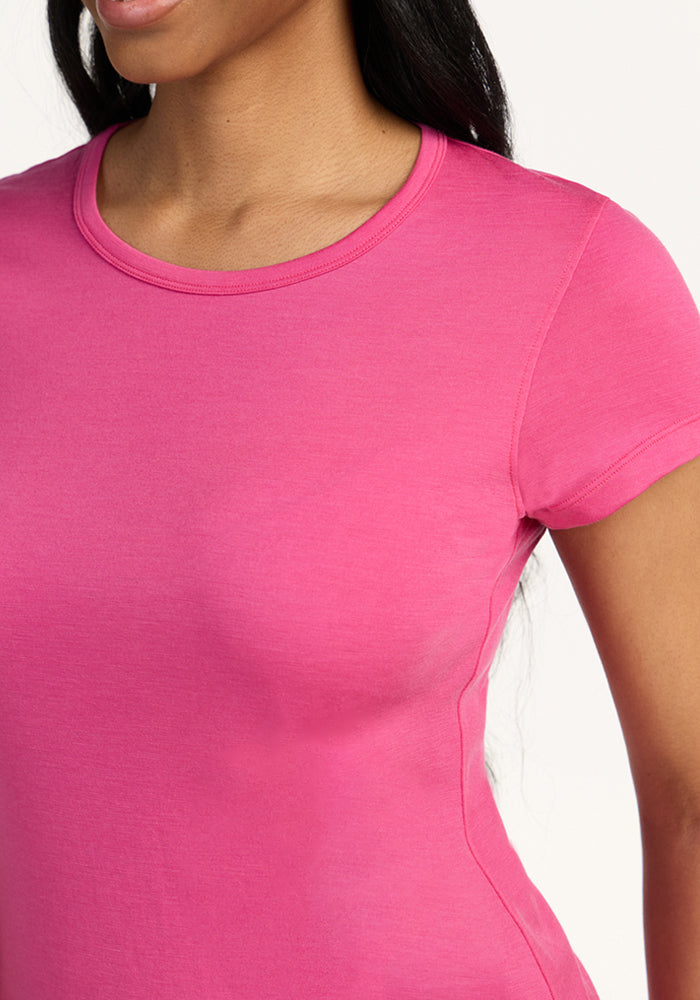 A woman wears the Woolx Addie Short Sleeve Crew in Raspberry Fizz, shown from shoulders to waist against a white background. The lightweight Merino wool keeps her comfortable and odor free. Her face is partly out of frame.