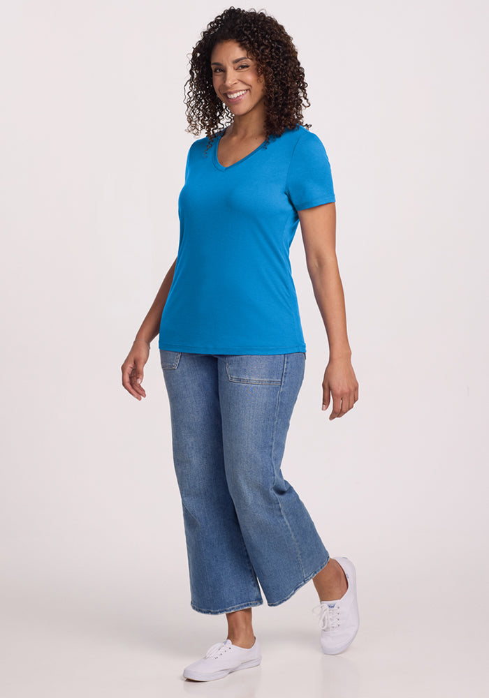 A woman with curly hair smiles while wearing the Woolx Junie V Neck - Wave Rider, paired with wide-leg blue jeans and white sneakers, standing against a plain light background.
