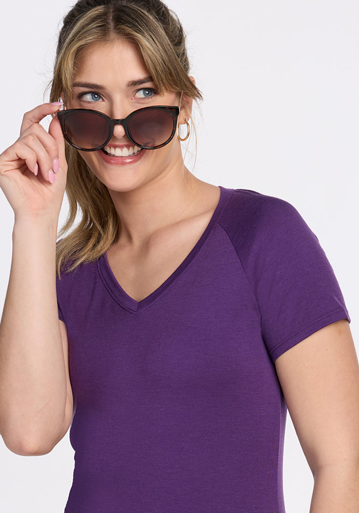 A smiling woman with light brown hair in a ponytail wears the Woolx Mia V Neck tee in Plum Crush and holds large sunglasses away from her face, looking to the side against a plain background.