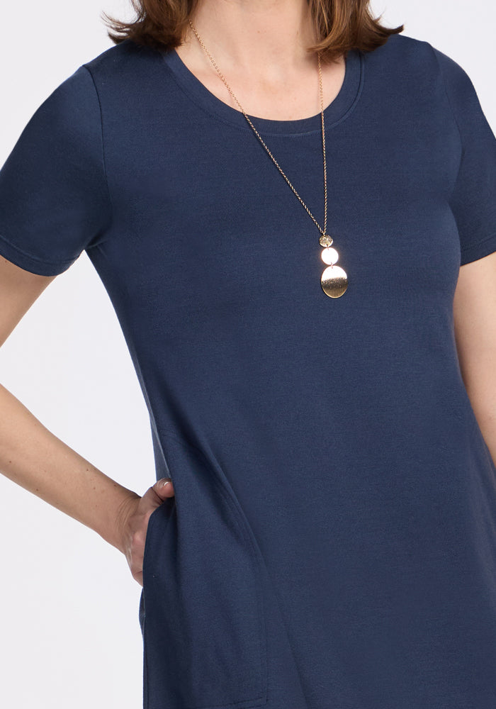 A woman with shoulder-length brown hair wears the Woolx Georgie Dress in Deep Navy. The Merino wool swing dress has short sleeves, pockets, and is paired with a gold pendant. Image is cropped at her neck and mid-thigh.