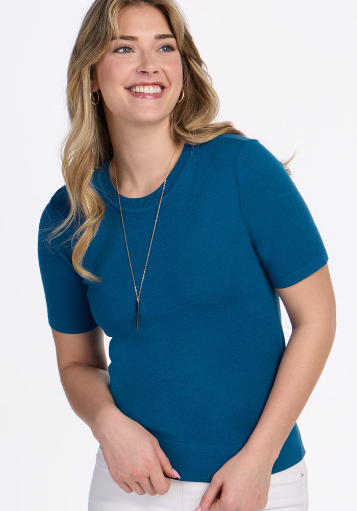 A smiling woman with long blonde hair wears the Woolx Anna Crew Sweater in Moroccan Blue, paired with a long necklace and white pants, standing against a plain white background.