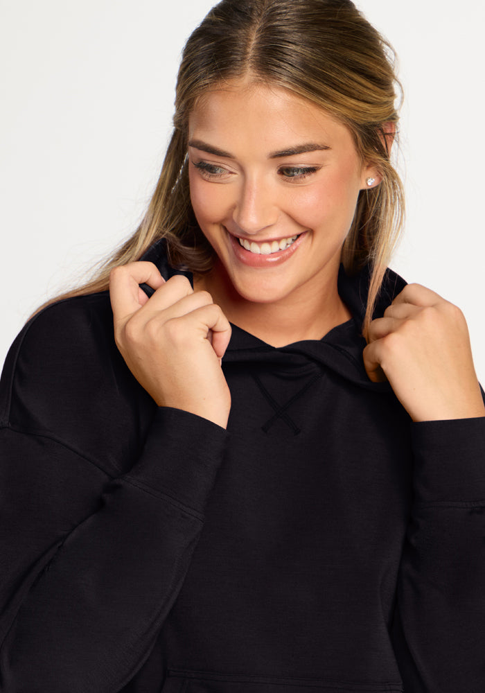 Smiling woman in the Woolx Lizzy Hoodie - Black holds the collar with both hands against a plain background.