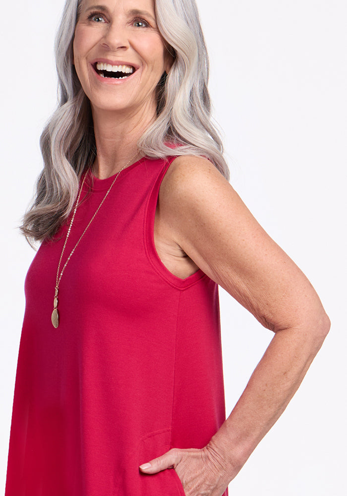 A smiling older woman with long gray hair wears the Woolx Clara Dress in Poppy Spark, a sleeveless, lightweight, breathable style, along with a long necklace and one hand in her pocket, standing against a plain white background.