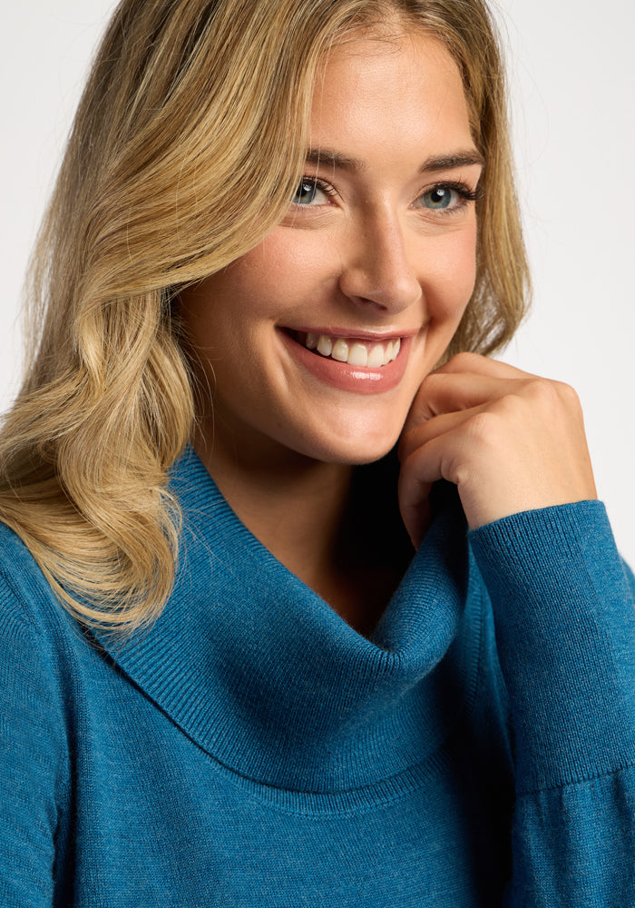 Smiling woman in Woolx Josie Cowl Neck Sweater—Blue Pine, blonde hair, gazing slightly off-camera.
