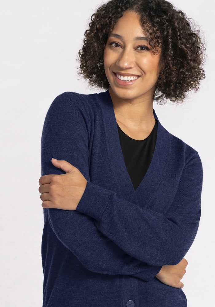 Woman with curly hair smiles, wearing Woolx Gia Sweater in Ocean Cavern over a black top, arms gently folded.