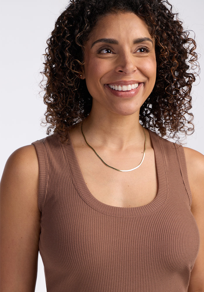 A woman with curly hair smiles while wearing the Woolx Sloane Ribbed Tank in Sienna Kiss and a thin gold necklace, standing against a plain light background.
