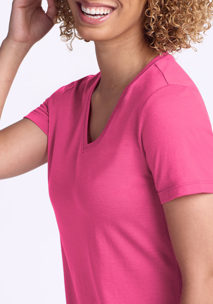 A person with curly hair and a bright smile wears the Woolx Junie V Neck in Raspberry Fizz. The short-sleeved, relaxed fit t-shirt is visible from the upper body only.