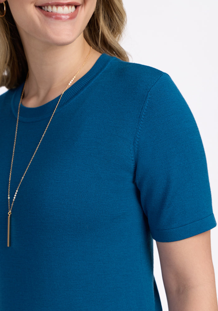 A woman smiles against a white background, wearing the Woolx Anna Crew Sweater in Moroccan Blue, paired with a long gold vertical bar necklace and gold hoop earrings.