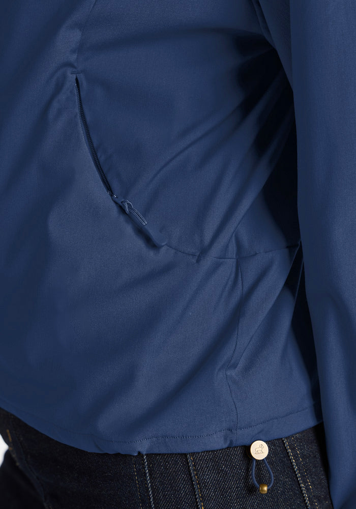 Close-up of a blue Cooper Windbreaker - Vintage Indigo showing a zippered side pocket and a metal logo tag at the hem.