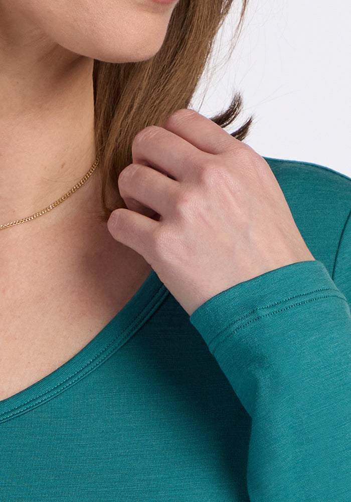 A close-up shows a person in the Woolx Layla V Neck Top - Surfside, touching their hair by their chin. A gold chain necklace is visible. The moisture-wicking, odor-resistant merino wool top is ideal for all-day comfort.