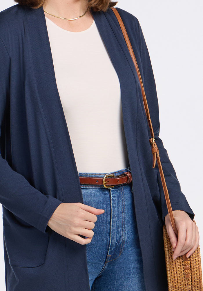 A person wears a Woolx Paisley Cardigan in Deep Navy, a white top, blue jeans with a brown belt, and carries a woven brown crossbody bag. Only the upper body is visible, with one hand tucked in a pocket.