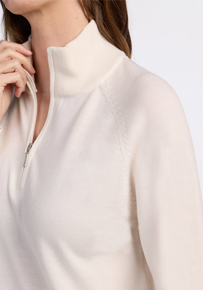 A close-up of a person wearing the Woolx Collins Half Zip Sweater in Dewdrop Pearl, featuring off-white merino wool, long sleeves, and a ribbed high collar, touching the collar with one hand against a plain light background.