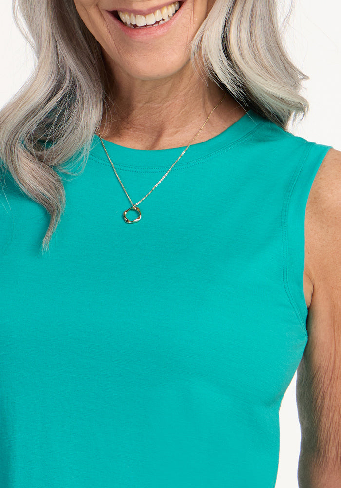 A smiling woman with long gray hair models the Woolx Bella Tunic Top in Cool Jade, paired with a gold round pendant necklace. Only her lower face, neck, and upper torso are shown against a white background.
