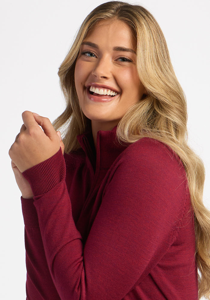 Woman with long blonde hair smiles in a Woolx Collins Half Zip Sweater in Beet Red against a white background.
