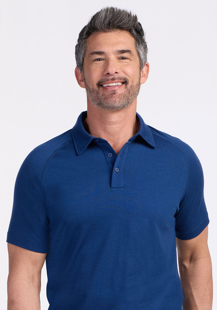 A smiling man with short gray hair and a trimmed beard is wearing the Woolx Summit Polo in Estate Blue, standing against a plain light background. The Merino wool fabric offers excellent moisture management.