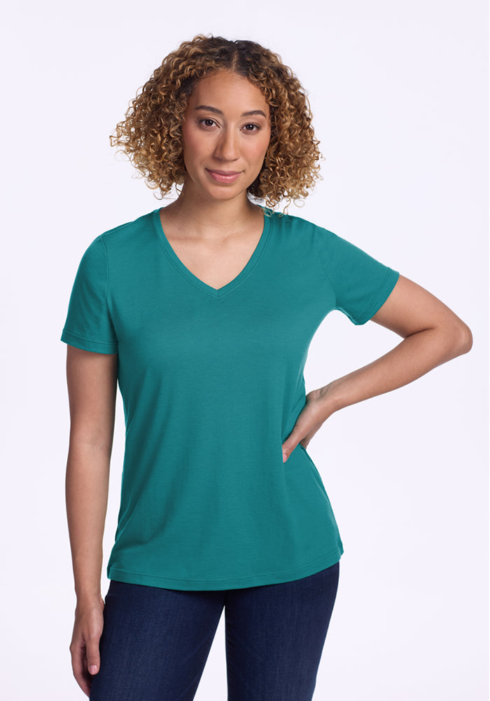 A person with curly hair is wearing the Woolx Junie V Neck - Surfside Merino wool tee in teal and dark jeans, standing with one hand on their hip against a plain white background. 