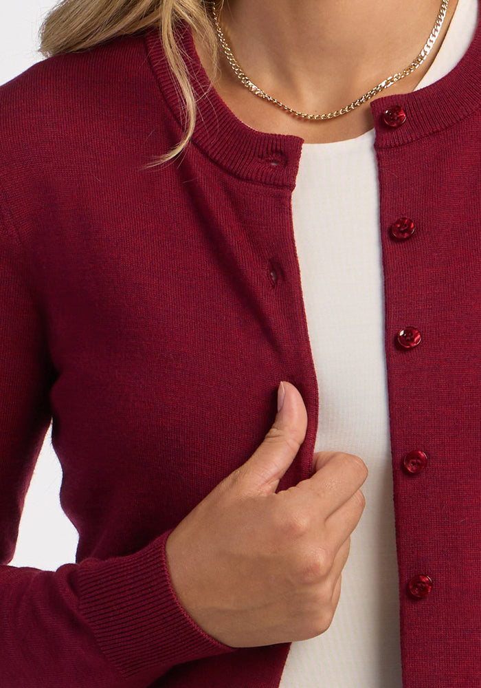 A woman in the Woolx Caitlin Sweater - Beet Red hugs it closed over a white top, accessorized with a gold chain necklace.