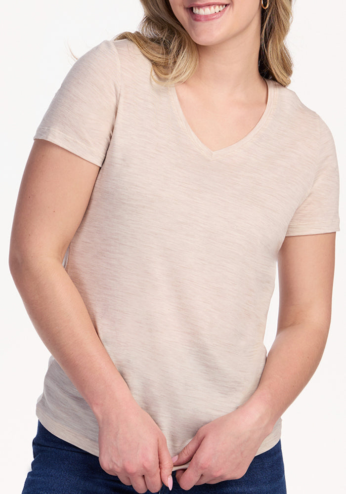 A woman wearing the Woolx Junie V Neck in Cream Heather and blue jeans stands against a white background, smiling with her hands loosely clasped in front of her.