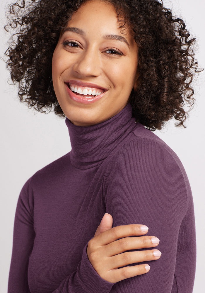 Curly-haired woman smiles warmly in the Woolx Sage Ribbed Turtleneck - Purple Iris, hand resting on her arm.