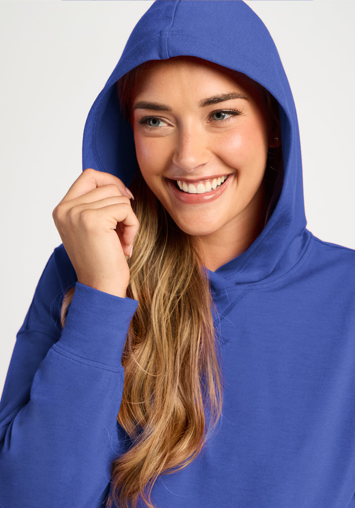 Smiling woman in a Woolx Lizzy Hoodie - Royal Blue, hood up, holding the edge and glancing to the side.