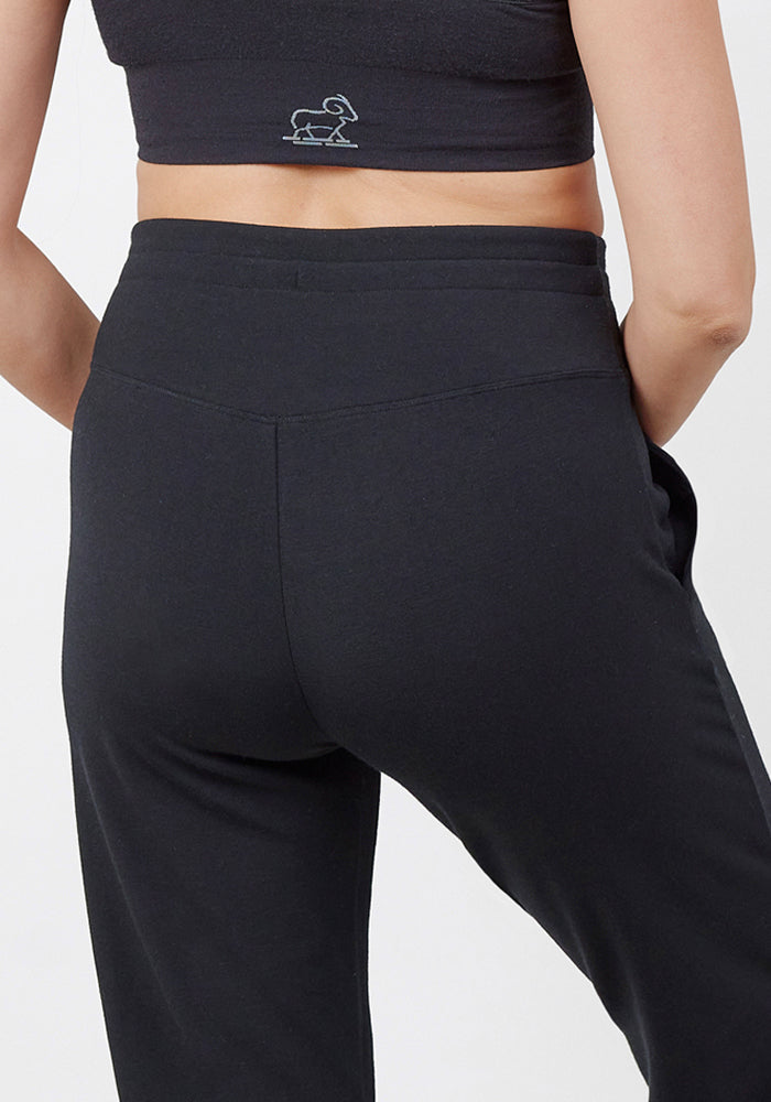 A person is shown from the back wearing Woolx Madden Joggers - Black and a black crop top with a small white embroidered dog graphic near the lower back, hands resting on hips.
