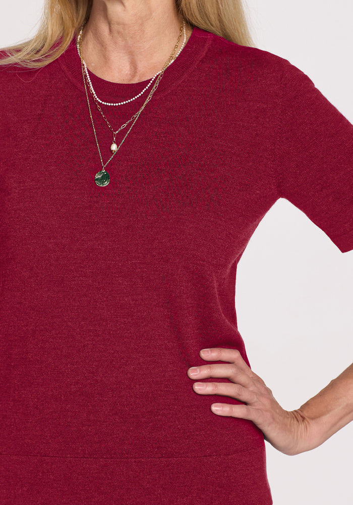 Woman wears Woolx Anna Crew Sweater in Beet Red, layered gold necklaces, hand on hip, white background.