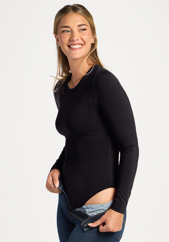 Smiling woman in the Woolx Margot Bodysuit - Black, pulling up blue jeans against a plain white background.