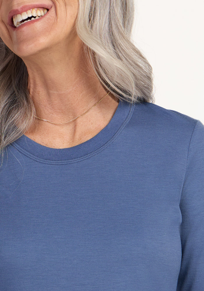 A smiling woman with long, wavy gray hair wears a simple gold necklace and the Woolx Hazel Tunic in Skyline, a blue crew-neck lightweight Merino wool top. The image shows her from the shoulders up, with her face partly out of frame.
