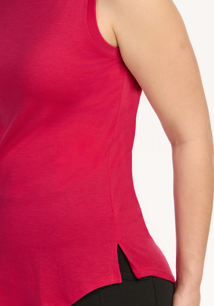 A close-up of a person wearing the Woolx Bella Tunic Top in Poppy Spark, a sleeveless women’s summer tank with a small side slit at the hem, paired with black pants. Only the torso and part of the arm are visible.
