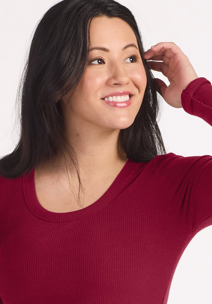 Woman wears the Woolx Caroline Ribbed Twirl Dress in Beet Red, smiling and touching her hair while looking to the side.