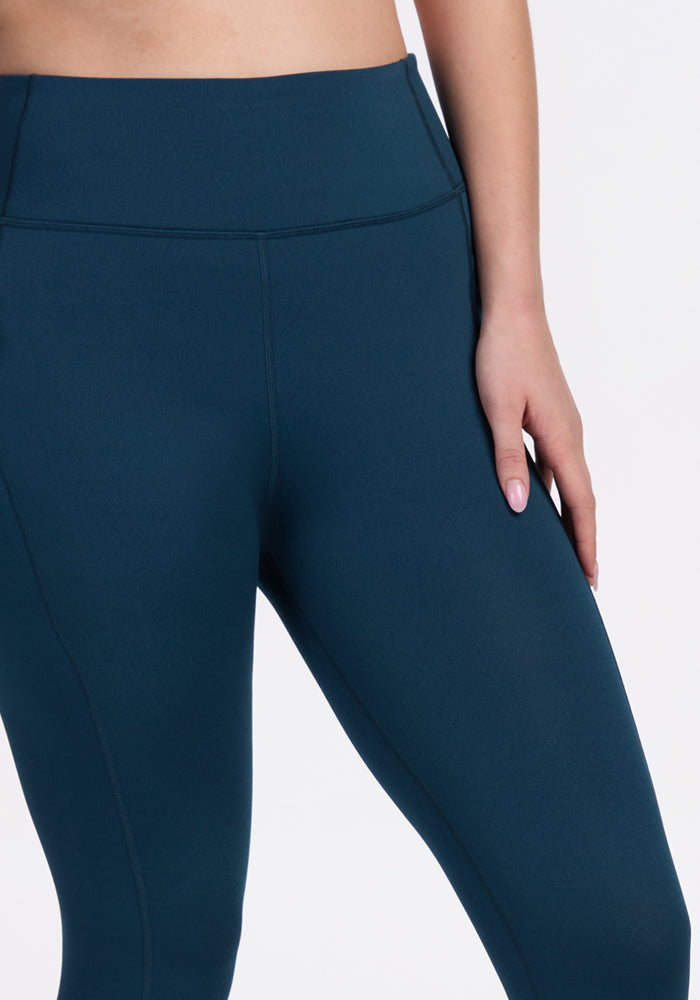 A person is shown from waist to mid-thigh wearing Woolx Merino Flex® McKenna Capris in Majolica Blue, with one hand at their side against a plain white background.