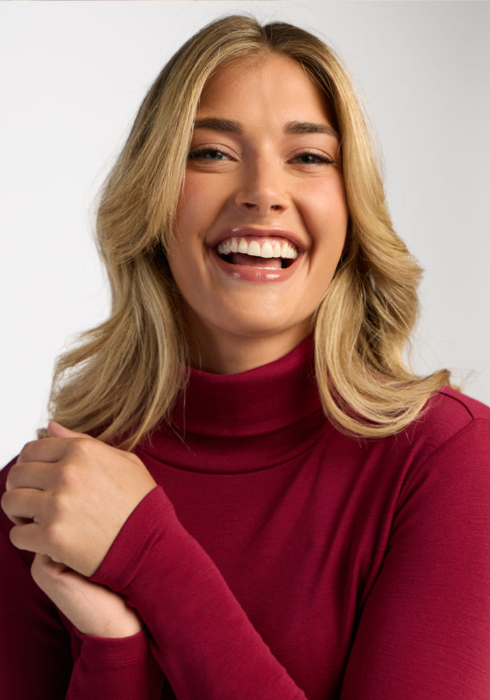 Smiling woman in a Woolx Peyton Turtleneck - Beet Red, blonde hair, hand near chest, against light background.