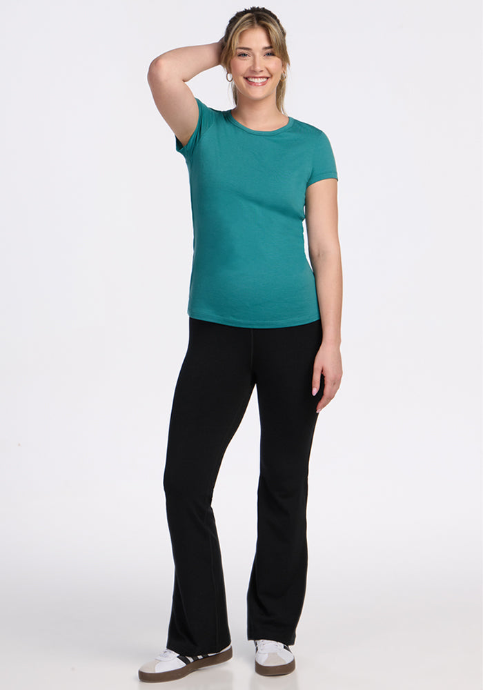 A smiling woman stands against a white background, wearing the Woolx Addie Crew Neck - Surfside, black pants, and white sneakers. One hand is on her head while the other rests by her side.
