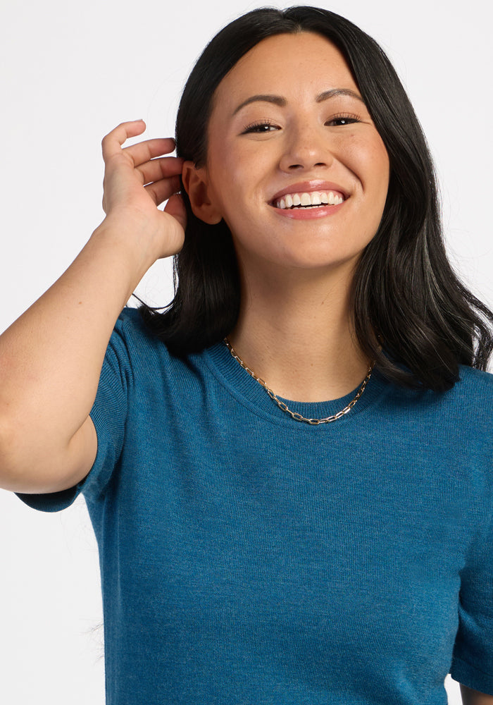 Smiling woman in a Woolx Anna Crew Sweater - Blue Pine, touching her ear and wearing a gold chain necklace.