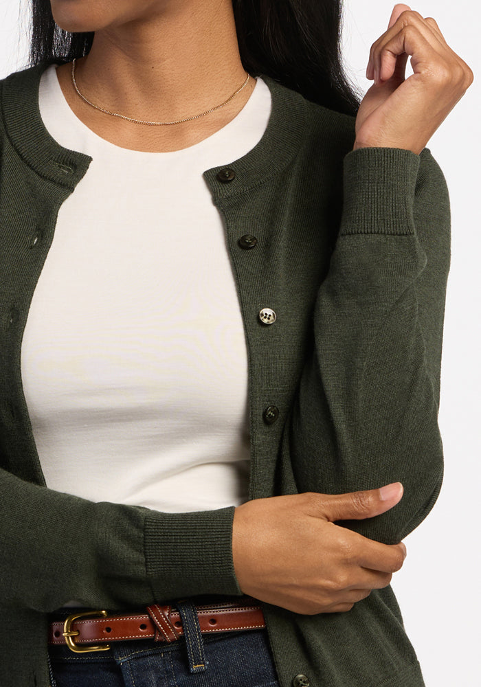Woman in a Caitlin Sweater - Duffel Bag by Woolx, white shirt, jeans with brown belt, holding arm and facing sideways.