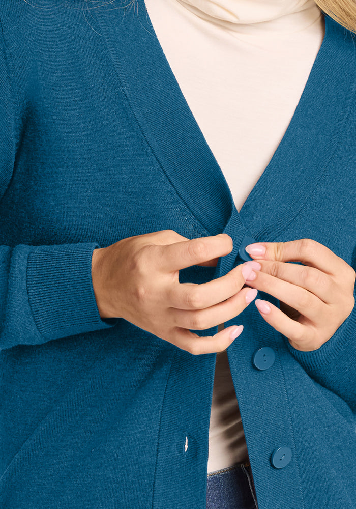 Person with light nail polish fastening a Gia Sweater - Blue Pine by Woolx over cream turtleneck, hands visible.