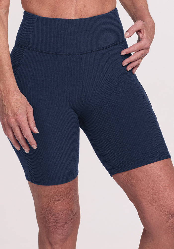 A person models the Woolx Miley Bike Shorts in Deep Navy, featuring a high-waisted ribbed design with pockets, standing with one hand on their hip against a plain light background.