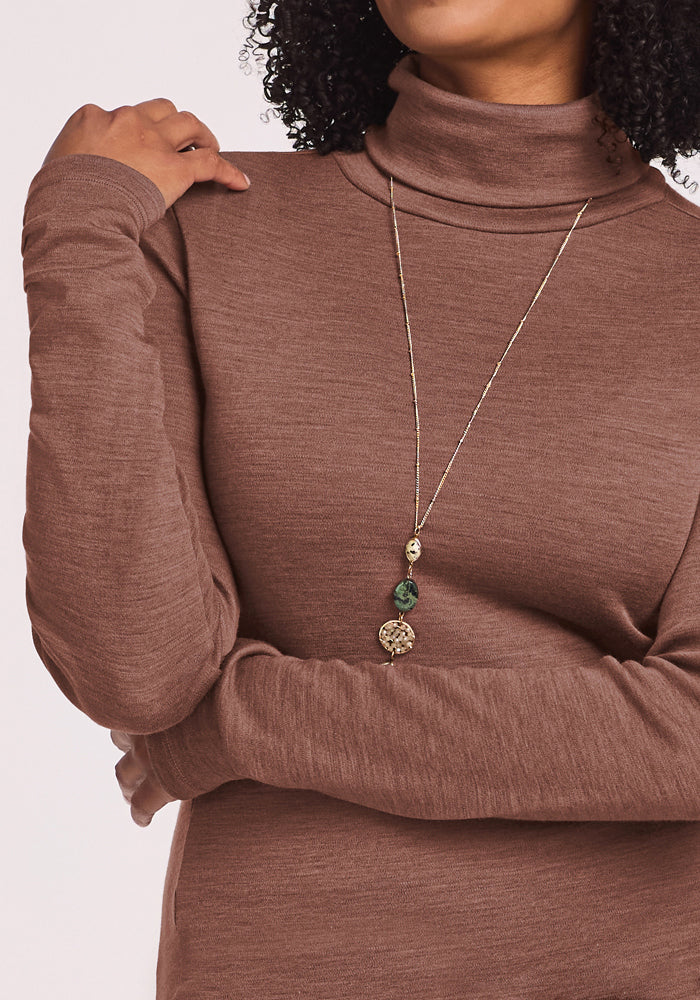 Person in Woolx Peyton Turtleneck - Sable Heather, wearing a long gold necklace with green and gold pendants.