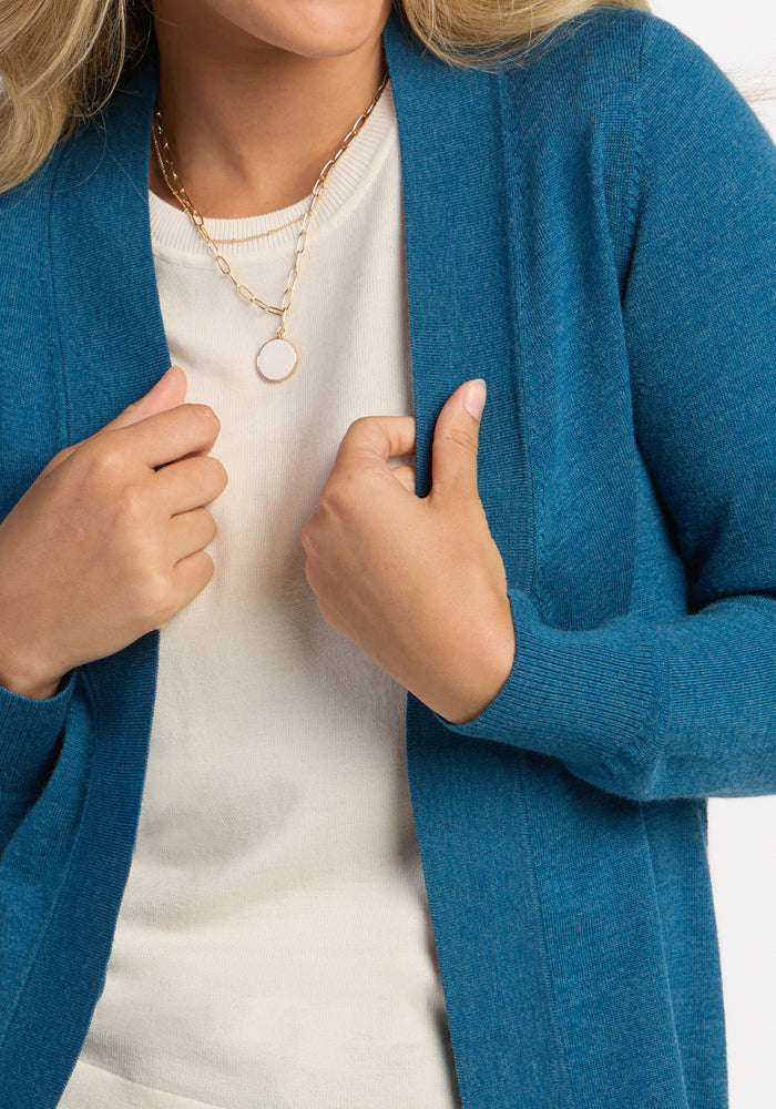 Woman in the Woolx Macie Sweater - Blue Pine and cream top, holding its edges; a gold necklace peeks through.
