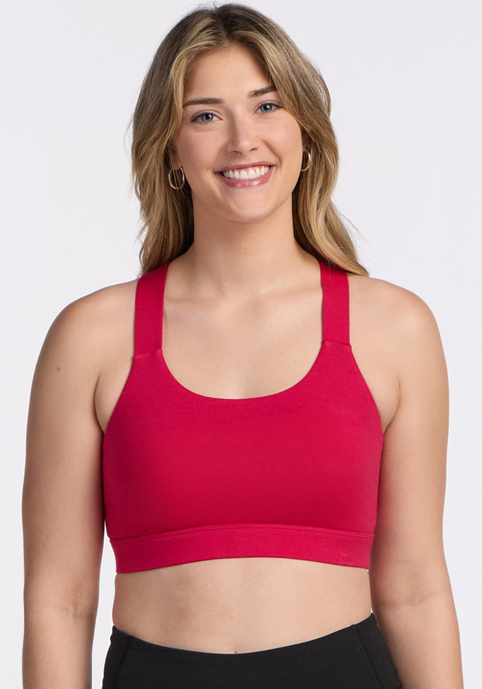 A young woman with long, light brown hair smiles at the camera, wearing the Woolx Livi Bra in Poppy Spark and black pants against a plain white background.