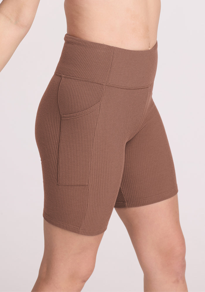 A person wearing Woolx Miley Bike Shorts in Sienna Kiss, featuring a high-waisted ribbed design with side pockets, is shown from the lower torso to upper legs against a light background.