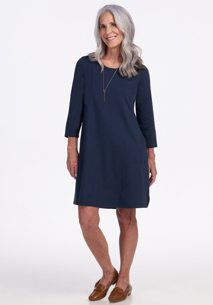 A smiling woman with long gray hair wears the Woolx Rissa A Line Dress in Deep Navy—a knee-length Merino wool dress with three-quarter sleeves—styled with a necklace and brown loafers, standing against a plain white background.