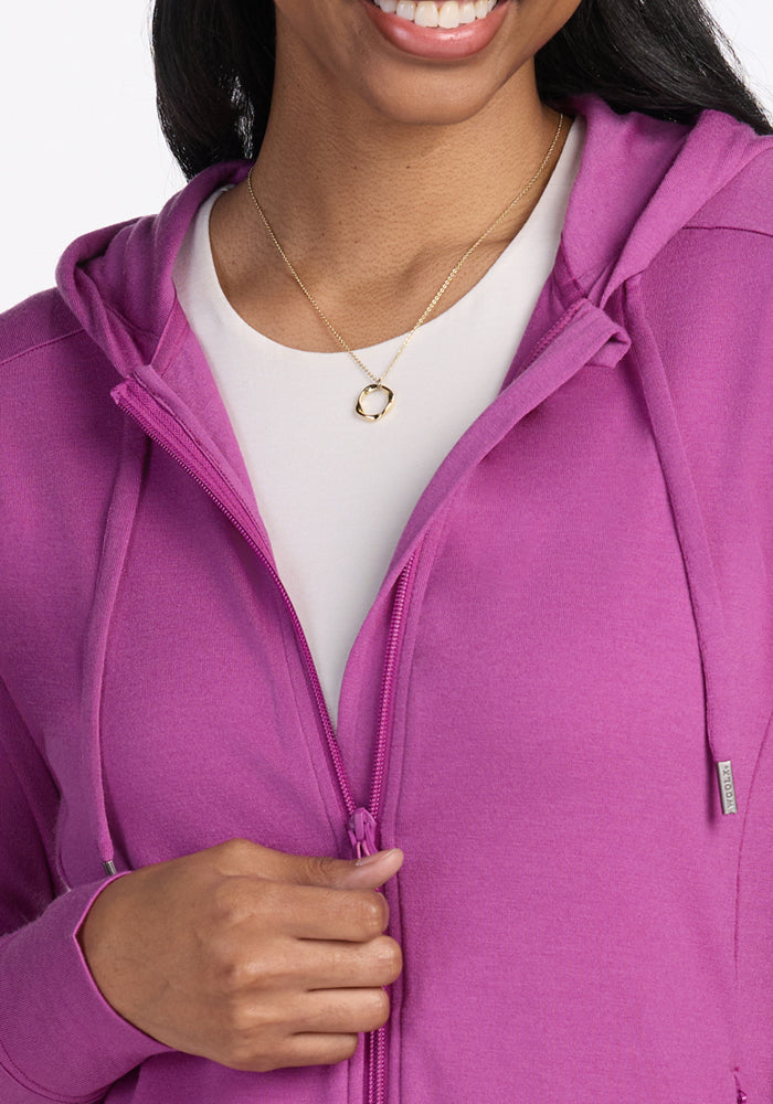 A woman smiles, holding the zipper of a Woolx Ryann Hoodie in Purple Wine over a white top, accessorized with a delicate gold necklace featuring a circular pendant.