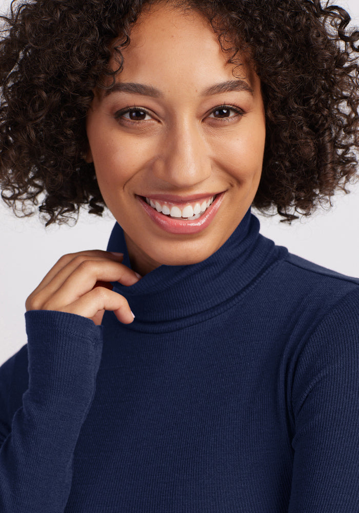 Smiling person with curly hair in a Woolx Sage Ribbed Turtleneck - Ocean Cavern, posing by a light background.